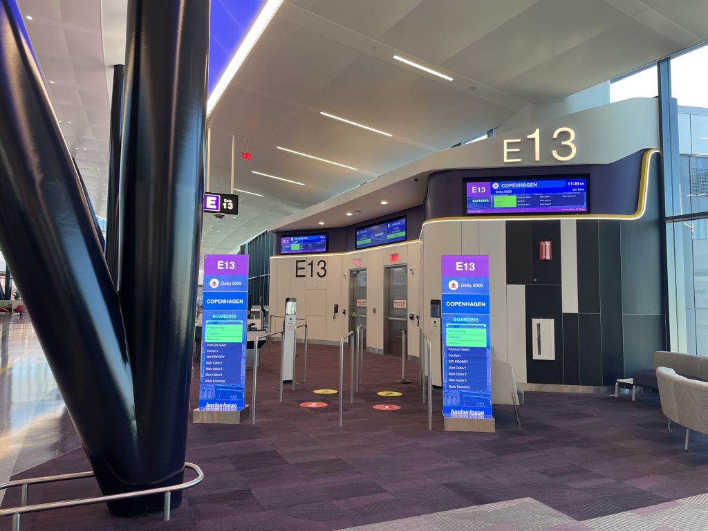 A wide view of 1/2 of gate 13 at Boston Logan Airport Terminal E Showing 1 Overhead horizontal Gate information Display and to LED totems in the foreground showing boarding information. 